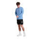 En mand står på en hvid baggrund iført en Saysky Clean Pace Long Sleeve, Herre, Blue, med sorte shorts, hvide sokker og hvide sneakers. Han kigger ned, mens han justerer det ene lange ærme.