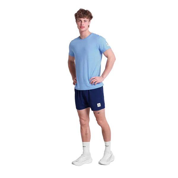 En mand står på en hvid baggrund iført en Saysky Clean Pace Running T-shirt, Herre, Blue, navy shorts, hvide sokker og sneakers. Med hænderne på hofterne og ansigtet vendt fremad viser han dette sporty herrelook.