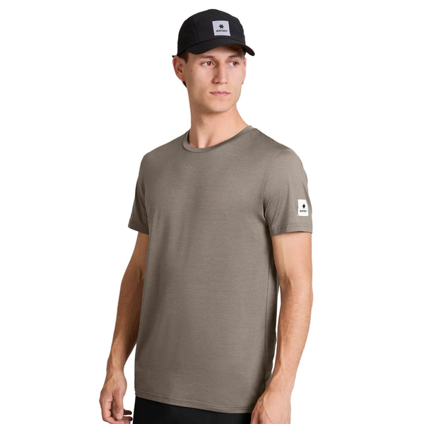 En mand iført en Saysky Clean Pace-T-shirt i taupe med et hvidt mærke på venstre ærme og en sort baseballkasket kigger til siden. Han står foran en ensfarvet hvid baggrund med højre arm afslappet ned langs siden.