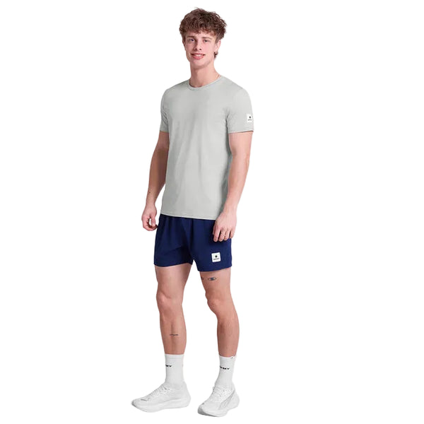 En mand ser frem for sig i en Saysky Clean Pace T-shirt, Herre, Grey, sammen med mørkeblå shorts, hvide strømper og hvide sneakers på en almindelig hvid baggrund.