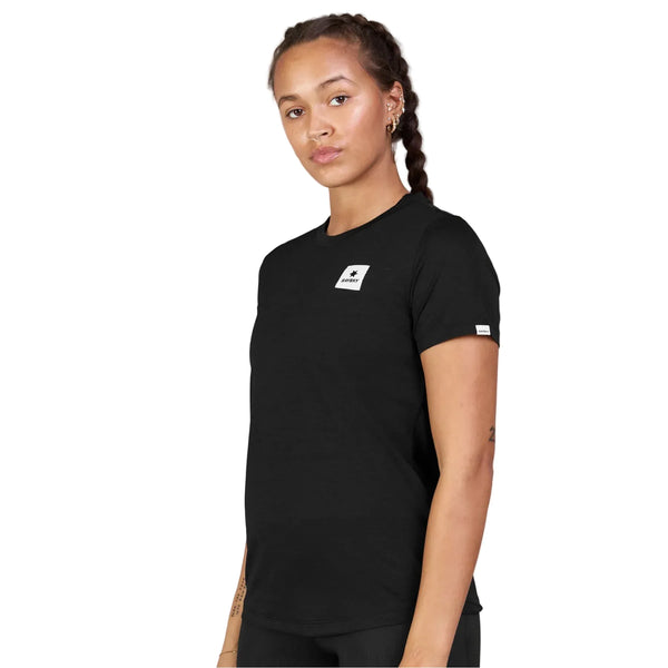 En kvinde med flettet hår har en Saysky Clean Combat Pro T-shirt til kvinder i sort på – en hurtigtørrende trænings-T-shirt med et lille hvidt Saysky-logo på brystet og en diskret mærkat på ærmet – mens hun står foran en hvid baggrund.