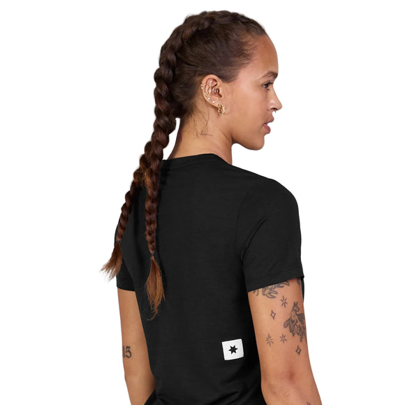 En kvinde med flettet hår, huller i ørerne og synlige tatoveringer på armene har en Saysky Clean Combat Pro T-shirt til kvinder i sort på, hvor der er en lille hvid firkant og en sort stjerne nederst på ryggen. Hun står med ryggen til kameraet, så man ser hendes profil.