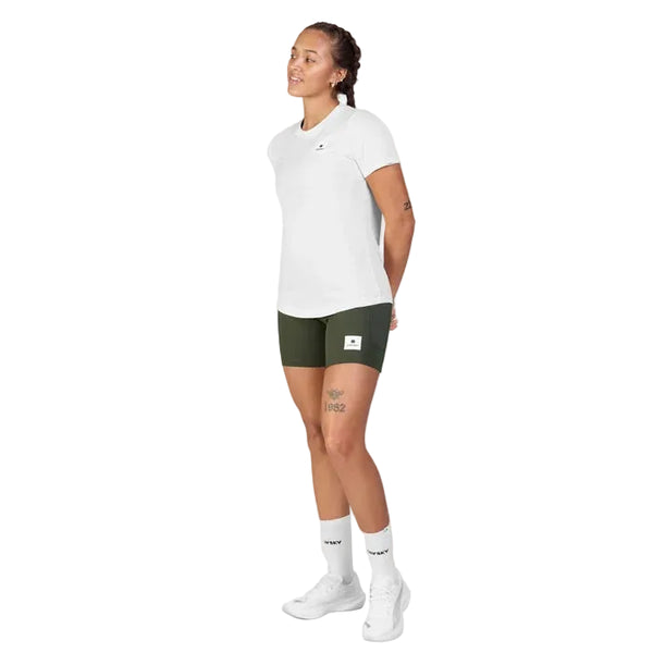 En person med flettet hår står vendt let mod venstre og er iført en hvid Saysky Clean Combat Pro T-shirt (Dame) sammen med mørkegrønne shorts, hvide sokker og hvide sneakers foran en ensfarvet hvid baggrund. Hænderne holdes bag ryggen. T-shirtens hurtigtørrende stof fra Saysky giver både komfort og funktionalitet til det sporty look.