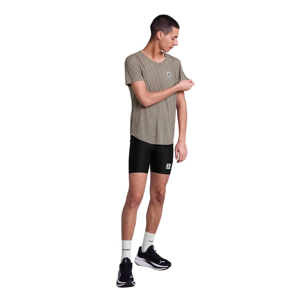 En mand står på en hvid baggrund iført en løs olivengrøn Saysky Reactive Flow T-shirt med mørkebeige logo, sorte sportsshorts, hvide sokker og sorte sneakers. Den fugttransporterende T-shirt holder ham afkølet, mens han kigger til siden og justerer sit ærme.