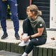 En kvinde i sportstøj med en grøn Saysky W Clean Pace T-shirt til kvinder sidder på en træplatform og smiler, mens hun binder sine hvide sneakers. I nærheden står en anden person i blå sportsbukser udendørs ved en bygning.