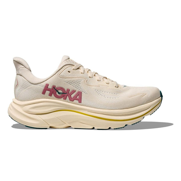 Hoka Clifton 10 Dame i Birch/Alabaster er en beigefarvet løbesko med tyk sål, overdel i mesh, pink "HOKA" på siden og gulgrønne detaljer på sålen. Den er let og komfortabel og vises, mens den kaster en skygge på en hvid baggrund.