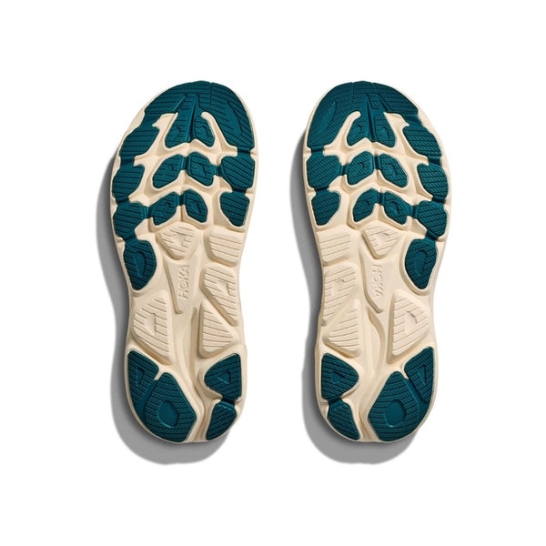 Et par Hoka Clifton 10 løbesko damesåler i Birch/Alabaster vises fra bunden, med en beige bund og blågrønt gummimønster for optimal støddæmpning. "HOKA"-branding er synlig på begge såler mod en hvid baggrund.