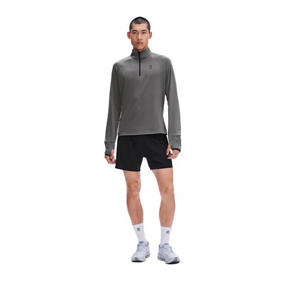 En person står med ansigtet vendt fremad, iført en grå On Climate Shirt 3, Herre, Ember, sorte shorts, hvide strømper og hvide løbesko foran en hvid baggrund – det perfekte tøj til løb i koldt vejr.