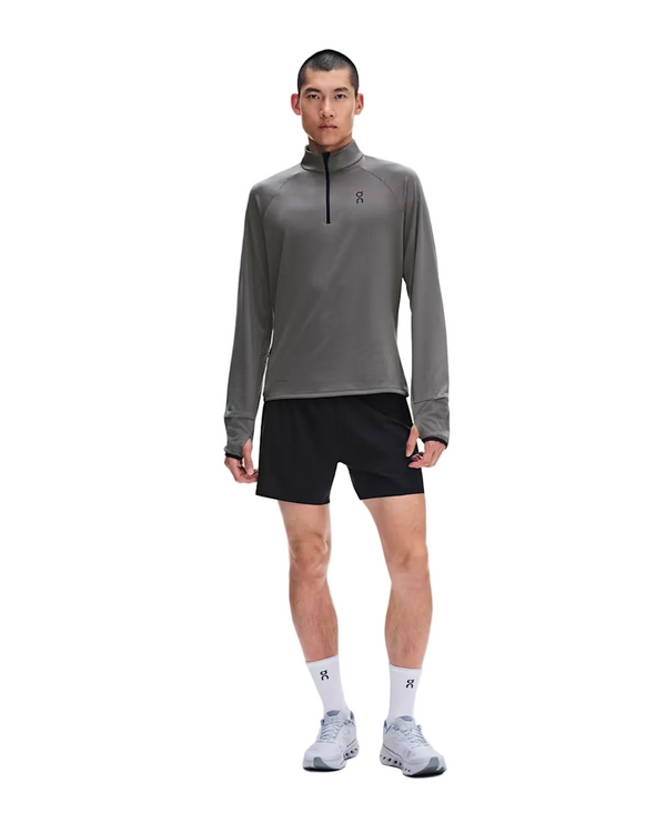 En person står med ansigtet vendt fremad, iført en grå On Climate Shirt 3, Herre, Ember, sorte shorts, hvide strømper og hvide løbesko foran en hvid baggrund – det perfekte tøj til løb i koldt vejr.