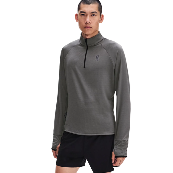 En person står med ansigtet fremad iført en grå On Climate Shirt 3, Herre, Ember langærmet løbetrøje med kvartlås og tommelfingerhuller samt sorte sportsshorts, foran en ensfarvet hvid baggrund.
