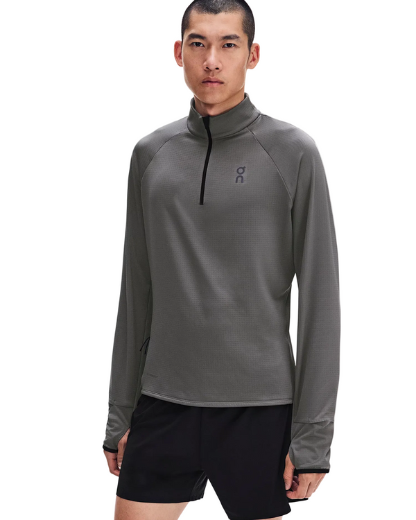 En person står med ansigtet fremad iført en grå On Climate Shirt 3, Herre, Ember langærmet løbetrøje med kvartlås og tommelfingerhuller samt sorte sportsshorts, foran en ensfarvet hvid baggrund.