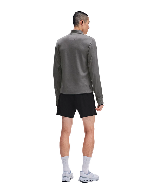 En mand står med ryggen til kameraet iført en grå On Climate Shirt 3, Herre, Ember – en perfekt løbetrøje til mænd i koldt vejr – sammen med sorte shorts, hvide sokker og grå løbesko. Han står foran en ensfarvet hvid baggrund.