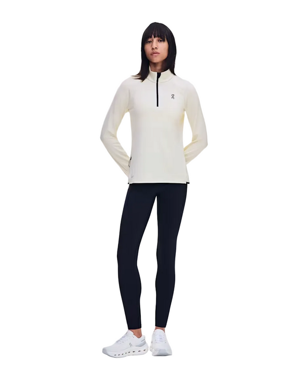En person står foran en hvid baggrund iført On Climate Shirt, Dame, Hare – en let langærmet løbetrøje med lynlås – kombineret med sorte leggings og hvide sneakers. Armene er afslappet bag ryggen, mens personen kigger fremad, klar til aktivitet selv i køligt vejr.