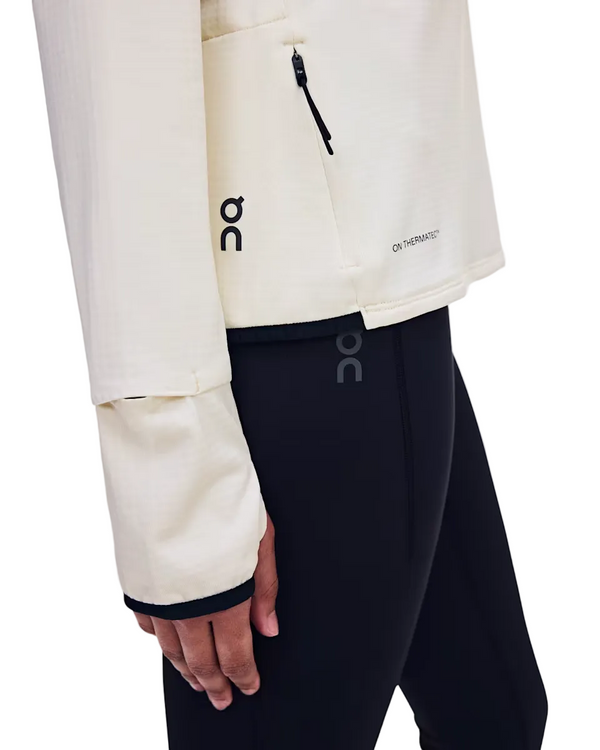 En person iført en lys On Climate-trøje, Dame, Hare-modellen med tommelfingerhuller og sorte leggings. Synlig lynlåslomme og On-logo på både trøjen og leggingsene. Denne trøje er ideel til køligt vejr. Kun den nedre del af overkroppen og armen er synlige.