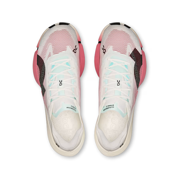 Oversigtsbillede af et par On Cloudboom Strike 2 unisex-sko i farven White/Camellia med lyserøde (camellia) og sorte detaljer, blå snørebånd og ON-logoet på tungen. Skoene er designet som konkurrencesko og er udstyret med et Carbon Speedboard for forbedret ydeevne. Den venstre sko er mærket med »CLOUDMONSTER STMM 6«. Parret vises side om side på en hvid baggrund.