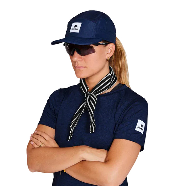 En person med langt lyst hår i hestehale bærer en Saysky Combat Cap, Unisex, Blue, mørke solbriller, en marineblå skjorte med ærmelap og et sort- og hvidstribet tørklæde - ideelt til løb takket være det hurtigtørrende stof - med krydsede arme mod en hvid baggrund.
