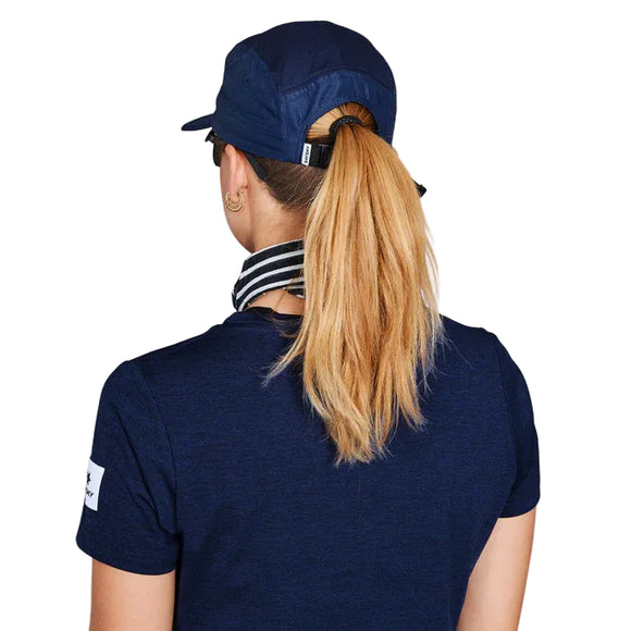 En person med langt lyst hår i hestehale ses bagfra iført Saysky Combat Cap Unisex Blue, en marineblå skjorte med stribet halsudskæring og en hvid patch på venstre ærme. Det hurtigtørrende outfit er ideelt til løb. Hvid baggrund.