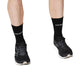En person er iført Saysky Combat High Socks, Unisex, Black, der er designet til performance-komfort. Kun deres underben og fødder er synlige, mens de står på en hvid baggrund i sorte løbesko.