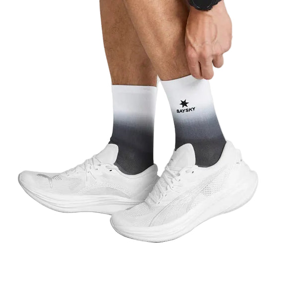 En person retter på den ene sok, mens vedkommende har Saysky Combat High Socks, unisex, sort/hvid, på sammen med hvide løbesko. Kun underbenene, sokkerne og skoene er synlige på en ensfarvet hvid baggrund, hvilket fremhæver den høje komfort, som disse træningssokker byder på.