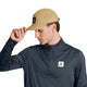 Personen, der bærer en beige Saysky Combat Shield-kasket og en marineblå unisex-langærmet trøje i funktionsmateriale med Saysky-logoet, retter kasketens skygge, mens vedkommende kigger ned mod venstre.