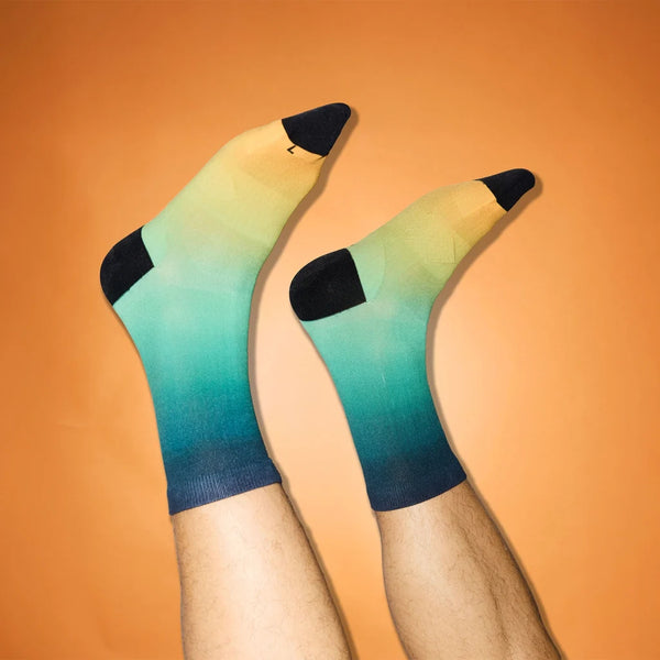 En person med lys hud løfter begge ben iført Saysky Drip Dye Combat Socks 200, Unisex, Blue Aop-gradient socks fading from yellow to teal with black toes, heels, and cuffs - set against a orange background.