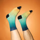 En person med lys hud løfter begge ben iført Saysky Drip Dye Combat Socks 200, Unisex, Blue Aop-gradient socks fading from yellow to teal with black toes, heels, and cuffs - set against a orange background.