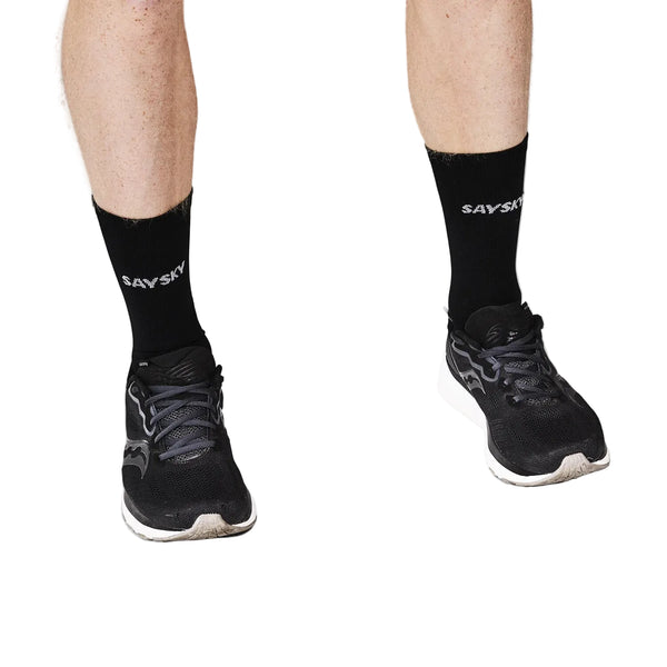 En person iført sorte Saysky Combat High Socks 100 og sorte løbesko står på en hvid baggrund, hvor kun underben og fødder er synlige.