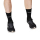 En person iført sorte Saysky Combat High Socks 100 og sorte løbesko står på en hvid baggrund, hvor kun underben og fødder er synlige.