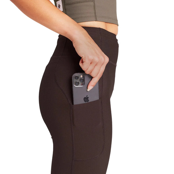 En person iført Saysky Combat+ Rib Long Tights, Dame, Coffee og en olivengrøn top lægger en sølvfarvet iPhone i sidelommen på leggingsene. Billedet er beskåret, så det kun viser overkroppen og overbenene.