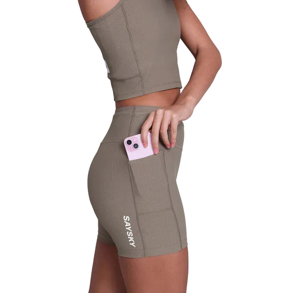 En kvinde i matchende taupe Combat+ Rib Running Short Tights 4" fra Saysky stikker en pink telefon ned i sidelommen; "SAYSKY" er trykt på shortsene, som er lavet af åndbar træningstøj.