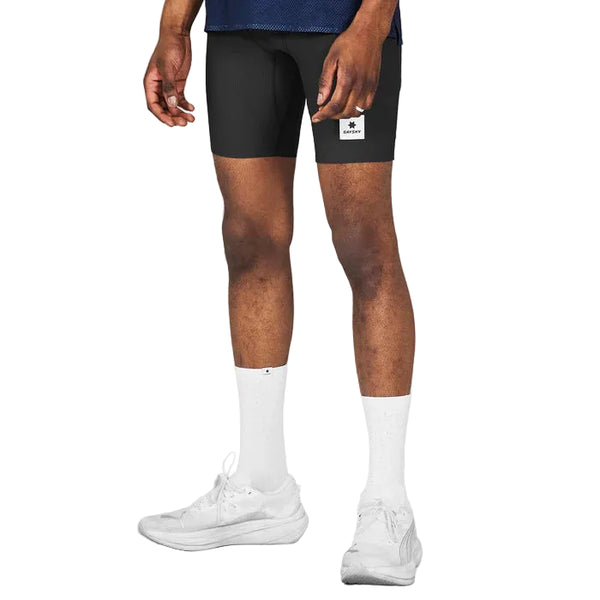Iført Saysky Combat+ Rib Running Short Tights 8, Herre, Black med en mørkeblå skjorte og hvide sneakers står en person med ansigtet vendt væk fra kameraet. Kun benene og underkroppen er synlige, hvilket understreger bevægelsesfrihed og åndbarhed.