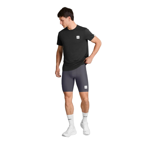 En mand står på en hvid baggrund iført en sort T-shirt, Saysky Combat+ Rib Short Tights 8″ Herre i gråt, hvide sokker, der går til læggen, og hvide sportssko. Han kigger til venstre og holder fast i bunden af sin T-shirt med den ene hånd, hvilket fremhæver de åndbare træningstights fra Saysky, der er perfekte til enhver træning.