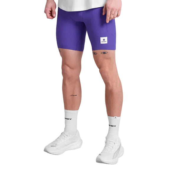 En person i hvid skjorte, Saysky Combat+ Rib Running Short Tights 8" Herre Purple, hvide sokker og sportssko står med ansigtet fremad. De tætsiddende shorts giver bevægelsesfrihed og åndbarhed. Tatoveringer er synlige på lårene og venstre underben.