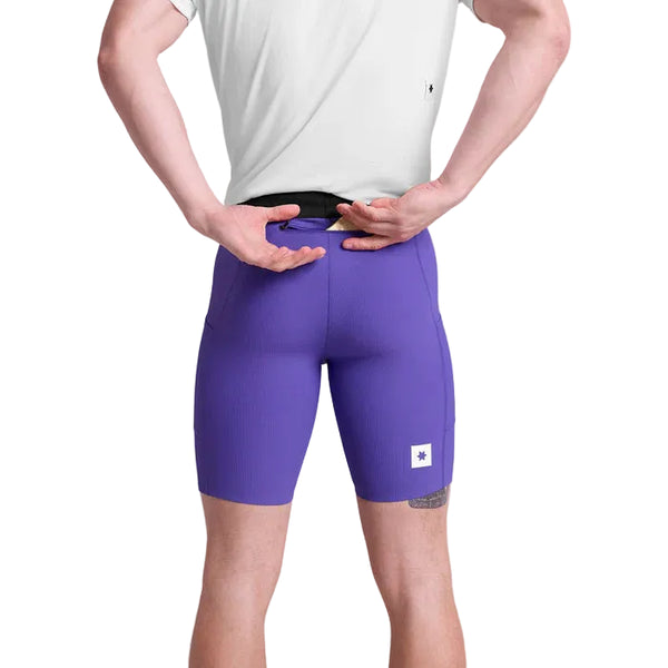 En person kigger væk fra kameraet og justerer linningen på Saysky Combat+ Rib Running Short Tights 8" til mænd i lilla med et lille hvidt mærke, der viser både åndbarhed og bevægelsesfrihed.