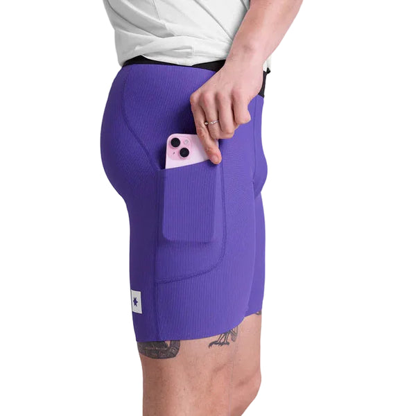 En person bærer Saysky Combat+ Rib Running Short Tights 8" til mænd i lilla og stikker en pink smartphone ned i sidelommen. Kun underkroppen, lårene, en del af en hvid skjorte og en tatovering er synlige.