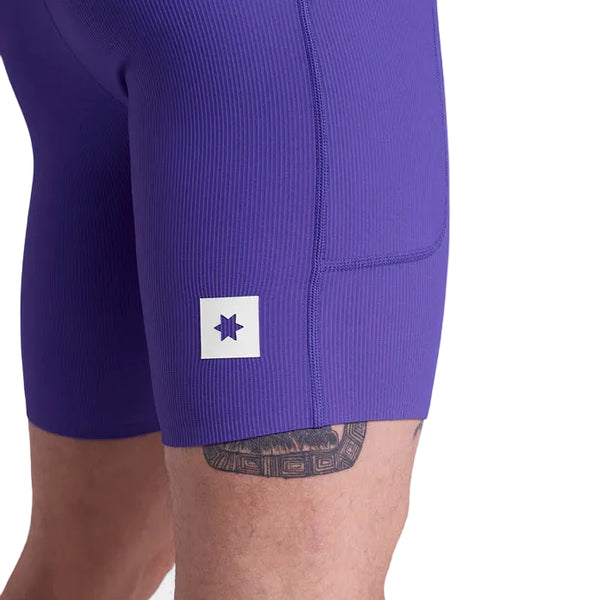 En mand er iført tætsiddende, ribbet lilla Saysky Combat+ Rib Running Short Tights 8" med et rektangulært hvidt mærke med et stjernelignende symbol på låret; en del af en tatovering på benet er synlig og fremhæver åndbarhed og bevægelsesfrihed.