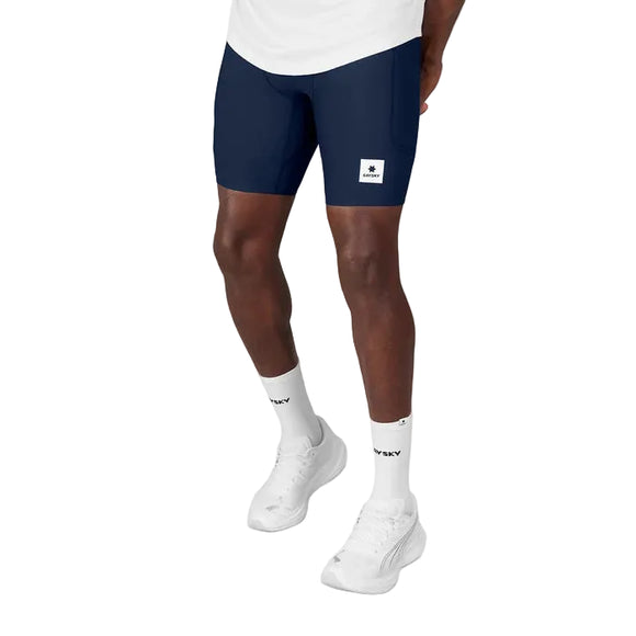 Der vises et billede af en person fra livet og ned på en hvid baggrund, iført de blå Saysky Combat+ Rib Short Tights 8″ til mænd. De slanke, tætsiddende sportsshorts har et lille logo-mærke og er designet til aktiv bevægelse. Outfitet fuldendes med hvide ankelstrømper og hvide sportssko.