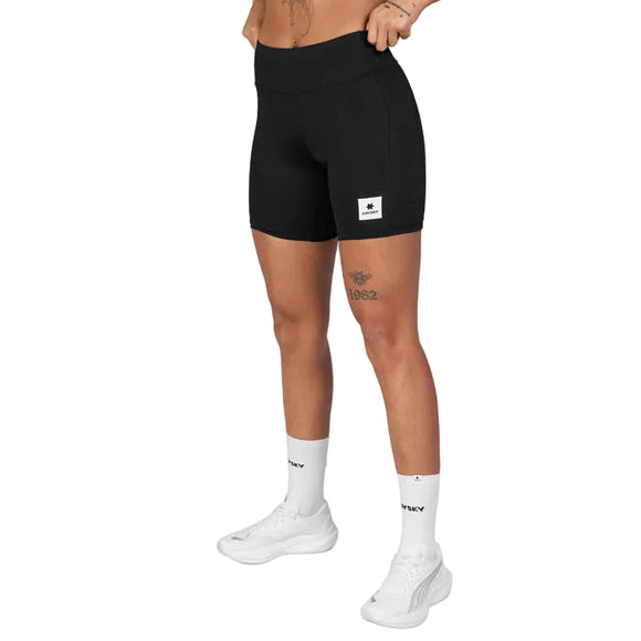 En person har Saysky Combat+ Short Tights 7″, Dame, i sort på – fremstillet af genanvendt polyester og med et påsyet logo – kombineret med hvide ankelstrømper og hvide sneakers, og står foran en ensfarvet lysegrå baggrund. Personens ansigt er ikke synligt på billedet.