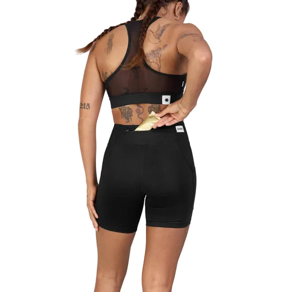 Man ser en person med tatoveringer bagfra, iført en sort sports-BH og Saysky Combat+ Short Tights 7″, Dame, i sort, fremstillet af genanvendt polyester. Personen lægger en guldfarvet genstand, muligvis en telefon eller en tegnebog, ned i baglommen på sine tights. Baggrunden er ensfarvet og lys.