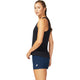 En kvinde står sidelæns mod en almindelig hvid baggrund iført en Asics Core Tank til kvinder i Performance Black - en svedtransporterende og hurtigtørrende top - og marineblå shorts med Asics-logo. Hendes hår er bundet tilbage i en fletning.