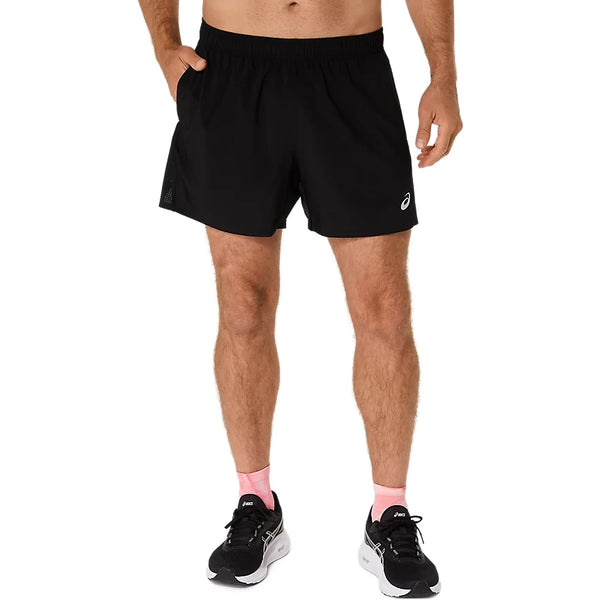 Der ses en person fra livet og ned på en hvid baggrund, iført ASICS Core 5In Shorts, Herre, Performance Black, en sort T-shirt, sort-hvide sportssko og lyserøde sokker.