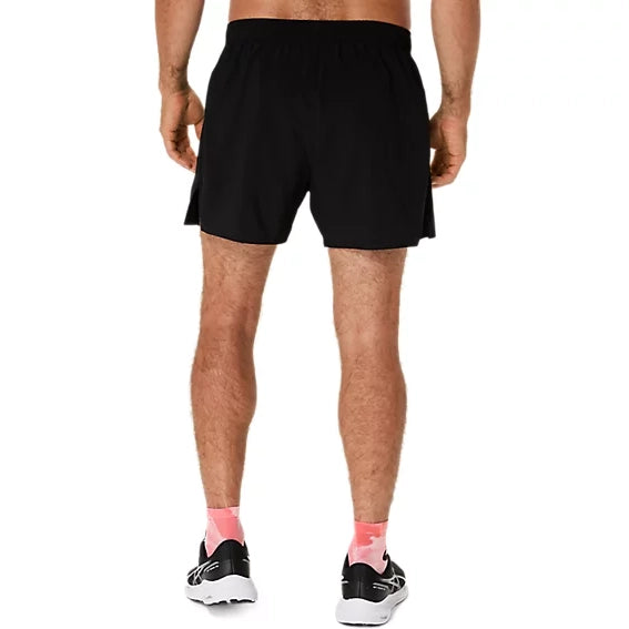 En mand står med ryggen til kameraet iført ASICS Core 5In Shorts Herre i farven Performance Black, sorte og hvide løbesko samt lyserøde sokker foran en ensfarvet hvid baggrund.