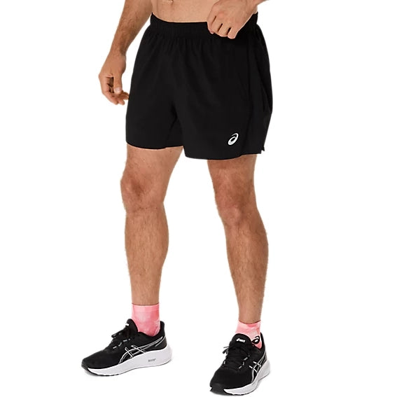 En person har Asics Core 5In Shorts, Herre, Performance Black på med et hvidt Asics-logo på venstre ben, sammen med sorte løbesko med hvide såler og lyserøde sokker. Overkroppen ses ikke på billedet.