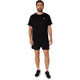 En mand står med ansigtet vendt fremad iført en sort T-shirt, Asics Core 5In Shorts Herre Performance Black, sorte sneakers med hvide såler og lyserøde sokker foran en ensfarvet hvid baggrund.