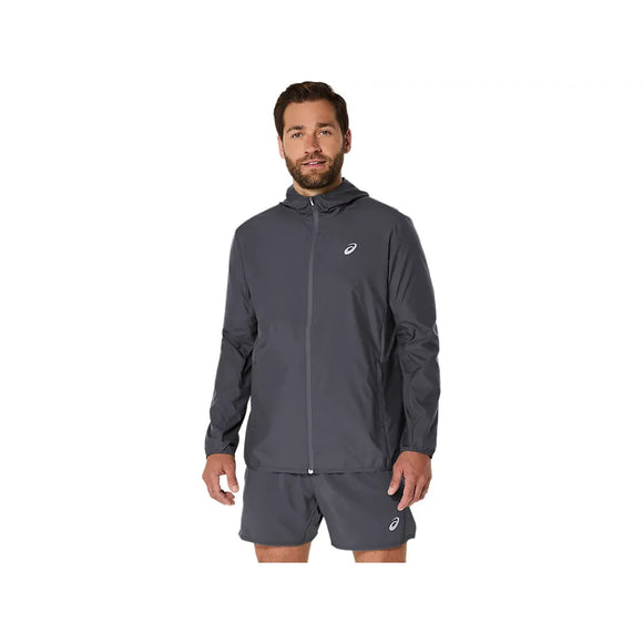 En mand med kort brunt hår og skæg har ASICS Core Jacket til mænd i farven Carrier Grey på – en vandafvisende jakke fra ASICS, der er designet med fokus på komfort. Han har den på sammen med et par matchende shorts og står med ansigtet vendt fremad, venstre arm let afslappet og højre hånd langs siden, foran en ensfarvet hvid baggrund.