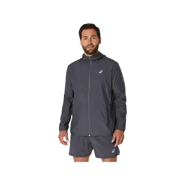 En mand med kort brunt hår og skæg har ASICS Core Jacket til mænd i farven Carrier Grey på – en vandafvisende jakke fra ASICS, der er designet med fokus på komfort. Han har den på sammen med et par matchende shorts og står med ansigtet vendt fremad, venstre arm let afslappet og højre hånd langs siden, foran en ensfarvet hvid baggrund.