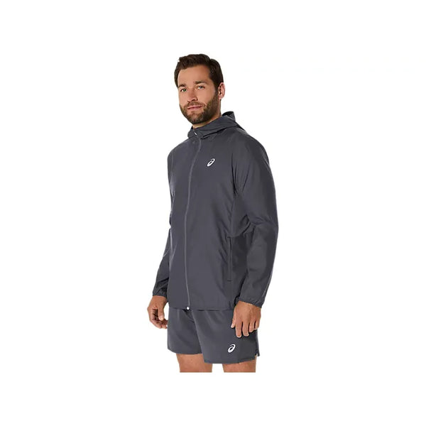 En mand har en Carrier Grey ASICS Core Jacket, Herre — en vandafvisende løbejakke til mænd — på sammen med et par matchende shorts. Begge dele har et lille hvidt ASICS-logo på venstre side. Jakken skiller sig ud mod den ensfarvede hvide baggrund, mens han kigger let til venstre.