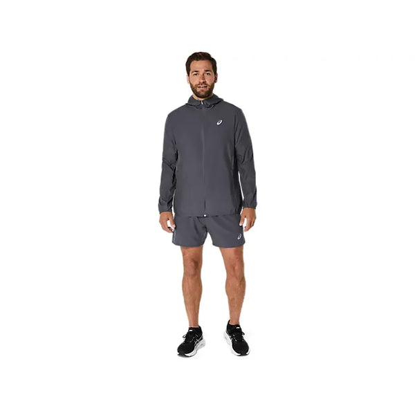 En mand står med ansigtet vendt fremad iført en grå Asics Core Jacket, Herre, Carrier Grey – en vandafvisende løbejakke til mænd, der er designet til løb – samt matchende grå shorts. Både jakken og shortsene har et lille hvidt Asics-logo. Han har desuden sorte løbesko med hvide såler på. Baggrunden er ensfarvet hvid.