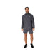 En mand står med ansigtet vendt fremad iført en grå Asics Core Jacket, Herre, Carrier Grey – en vandafvisende løbejakke til mænd, der er designet til løb – samt matchende grå shorts. Både jakken og shortsene har et lille hvidt Asics-logo. Han har desuden sorte løbesko med hvide såler på. Baggrunden er ensfarvet hvid.