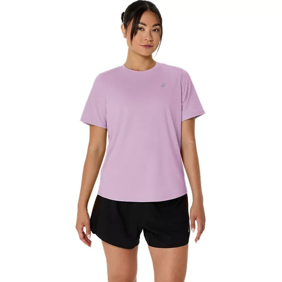 En person står med ansigtet vendt fremad iført en Asics Core Ss Top, Dame i farven Hazy Lilac med refleksdetaljer og sorte shorts, foran en ensfarvet hvid baggrund.